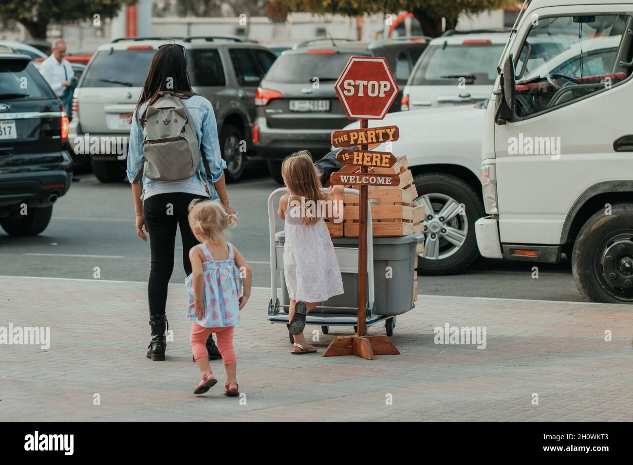 Mama mit Rucksack und Kindern auf dem Parkplatz auf der Suche und wartet auf ein Auto, um ihr Gepäck und Gepäck mit vielen Fahrzeugen im Hintergrund abholen Stockfoto