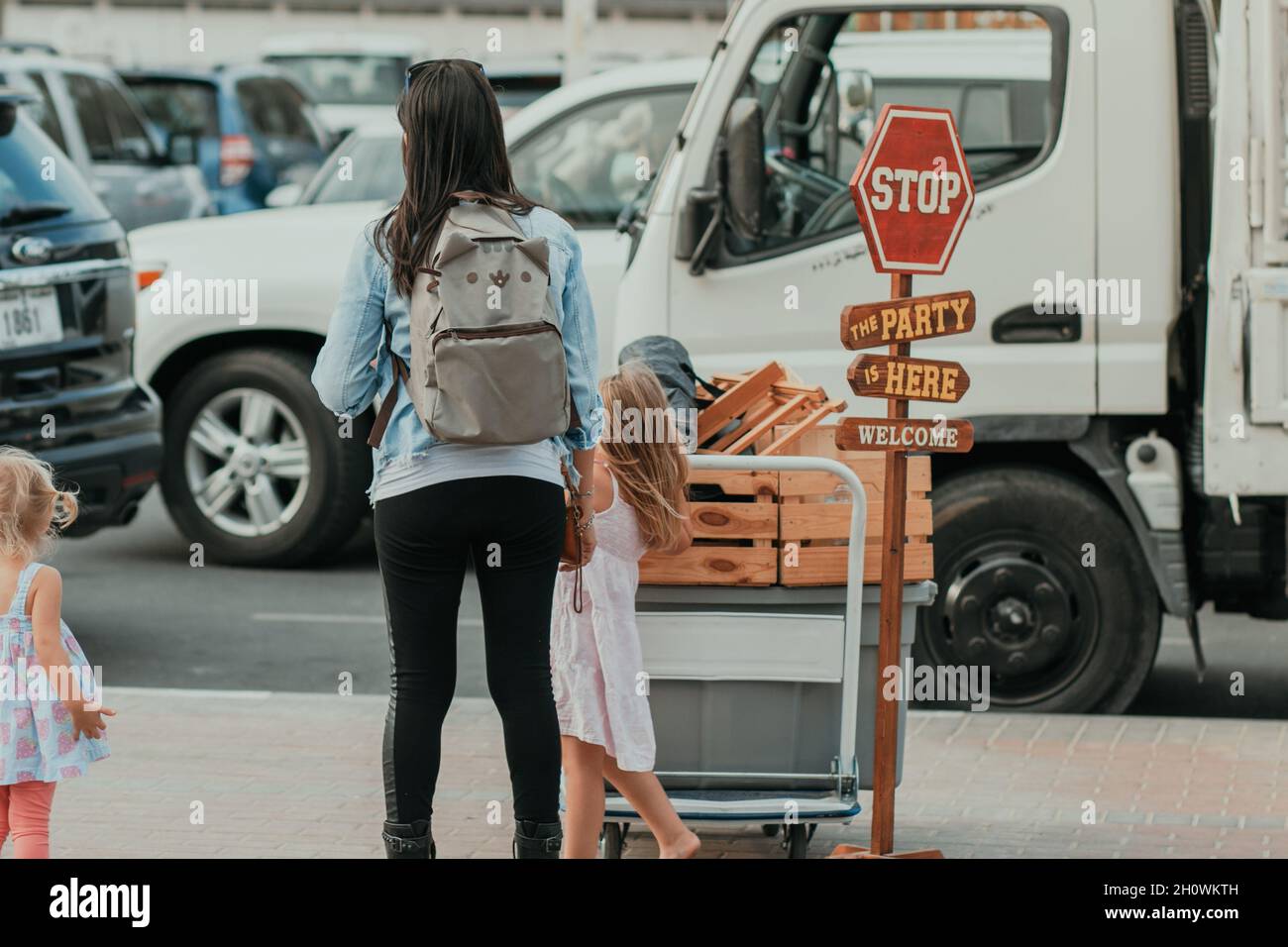 Mama mit Rucksack und Kindern auf dem Parkplatz auf der Suche und wartet auf ein Auto, um ihr Gepäck und Gepäck mit vielen Fahrzeugen im Hintergrund abholen Stockfoto