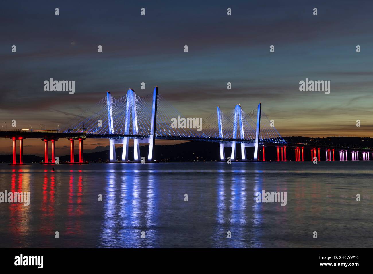 Die Governor Mario M. Cuomo Bridge, in Rot, Weiß und Blau zur Anerkennung des Columbus Day beleuchtet, überspannt den Hudson River kurz nach Sonnenuntergang. Stockfoto