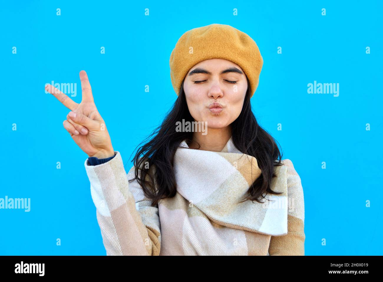 Foto von schönen verträumt Dame Schlag Luft Kuss zeigen V-Zeichen schließen Augen tragen beige Baskenmütze und Mantel isoliert Biue Farbe Hintergrund Stockfoto