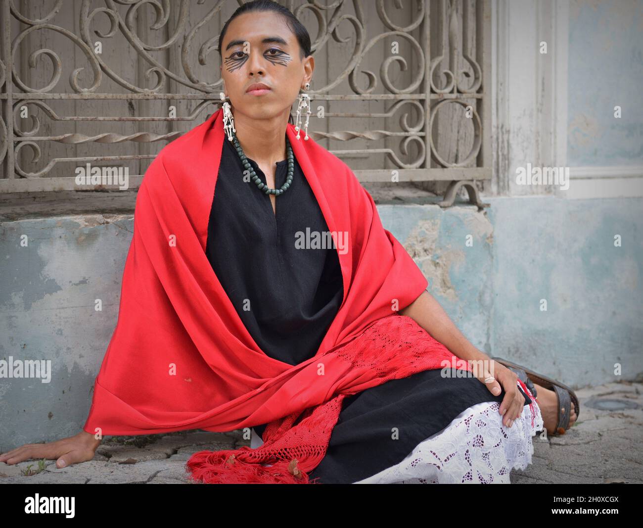 Der hübsche junge, nicht binäre Latino trägt einen roten mexikanischen rebozo und setzt sich auf den Bürgersteig vor einem traditionellen schmiedeeisernen Fenstergrill. Stockfoto