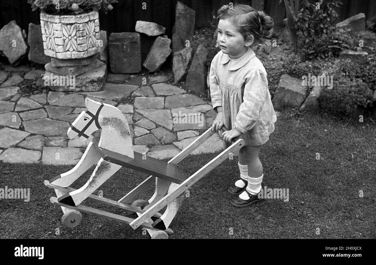 1960s, historisch, draußen auf einem Gras bei einer kleinen Terrasse, ein kleines Mädchen stehend, den Griff ihres Reitspielzeugs haltend, ein hölzerner Stoß entlang Pferd, England, Großbritannien. Stockfoto