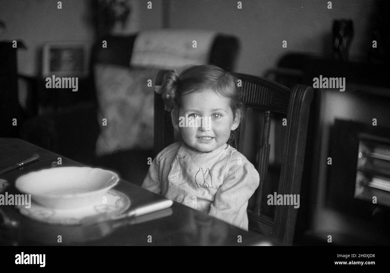 1950er Jahre, historisch, Teezeit und in einem Esszimmer, ein süßes kleines Mädchen, das auf einem Holzstuhl an einem formellen Esstisch sitzt, mit einer leeren Schüssel vor ihr, England, Großbritannien. Stockfoto