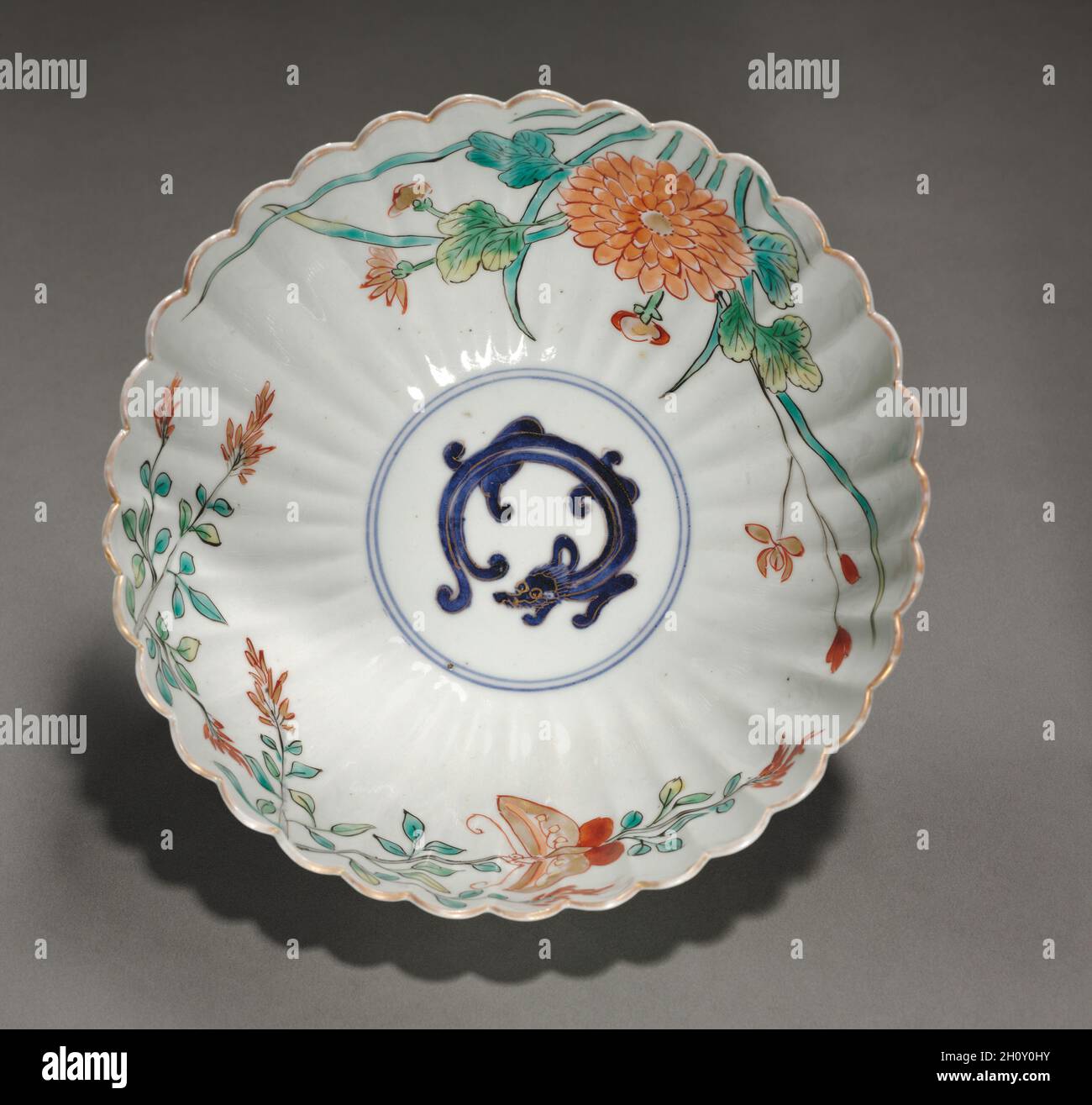 Geriffelte Schüssel mit Drachen, Schmetterling und Blumen, Anfang des 18. Jahrhunderts. Japan, Edo-Zeit (1615-1868). Porzellan mit Unterglasur in Blau und Überglasur in Emaille und Gold (Hizen Ware, Typ Kakiemon); Durchmesser: 18.6 cm (7 5/16 in.); Höhe: 8.6 cm (3 3/8 in.). Ein Drache, der von Blütenprays umgeben ist, und ein Schmetterling beleben das Innere dieser geriffelten Schale. Das Äußere hat eine ähnliche Dekoration, mit einem Hauch von Gold auf einigen Blütenblättern. Die Flöten verbessern sowohl das visuelle als auch das taktile Erlebnis; ihre konischen Formen ziehen das Auge von den Rändern nach innen und bieten Kanten für die Hände. Stockfoto