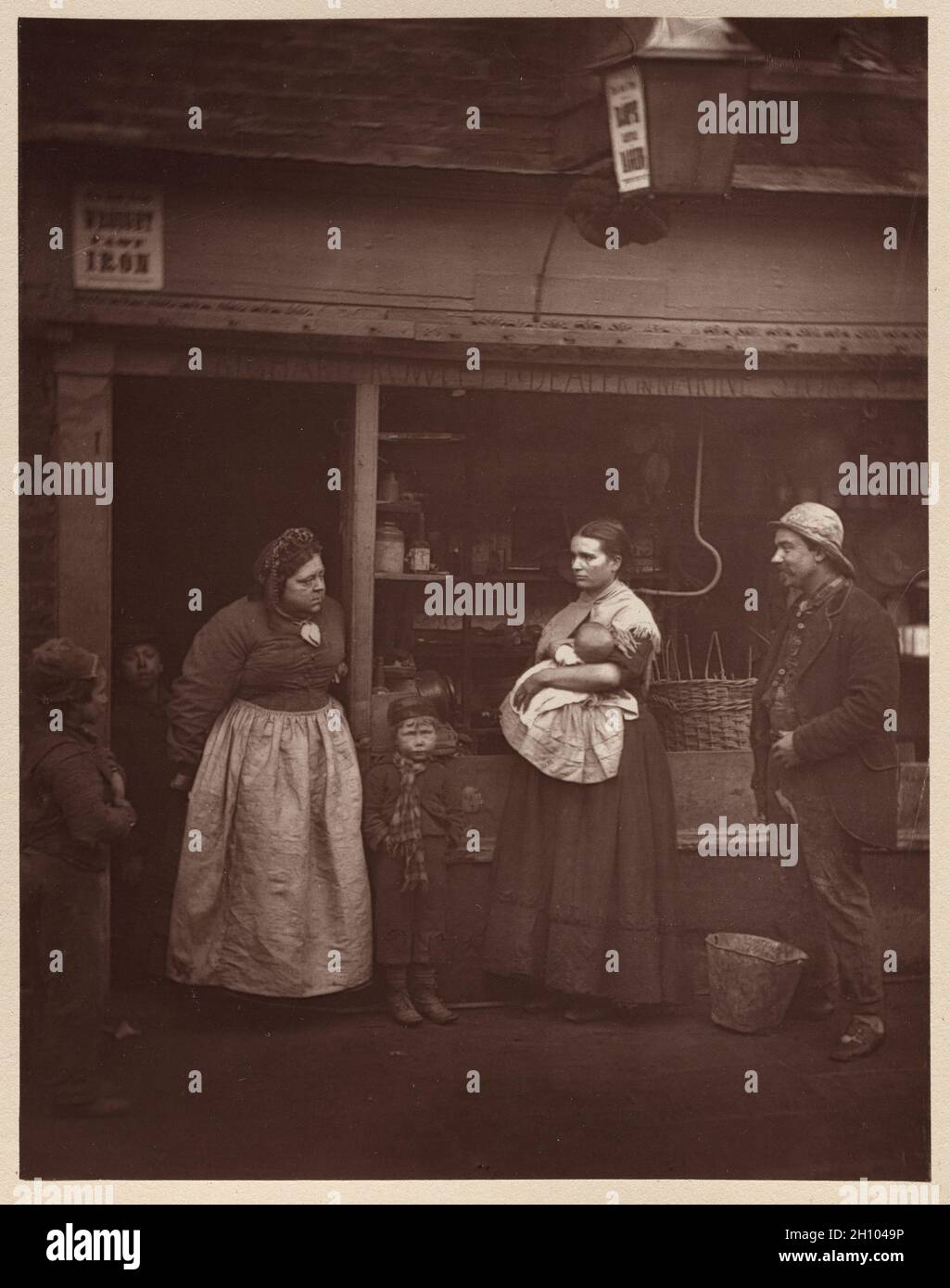 Street Life in London: Flutkranke, 1877. John Thomson (Scottish, 1837-1921). Woodburytype; Bild: 11.5 x 9 cm (4 1/2 x 3 9/16 Zoll); Papier: 11.5 x 9 cm (4 1/2 x 3 9/16 Zoll); montiert: 27 x 20.7 cm (10 5/8 x 8 1/8 Zoll). Smith und Thomson beschlossen, die einst wohlhabenden Rowletts vor dem ihnen eigenen und dort lebenden Schrottgeschäft zu fotografieren, um zu zeigen, dass die Überschwemmungen langfristige finanzielle Verluste und Gesundheitsprobleme verursachten, die selbst die Mittelschicht in die Armut trieben. Die Frau mit dem Baby wohnte im Haus nebenan; ihre ganze Familie litt unter ständigen Erkältungen und Rheuma Stockfoto