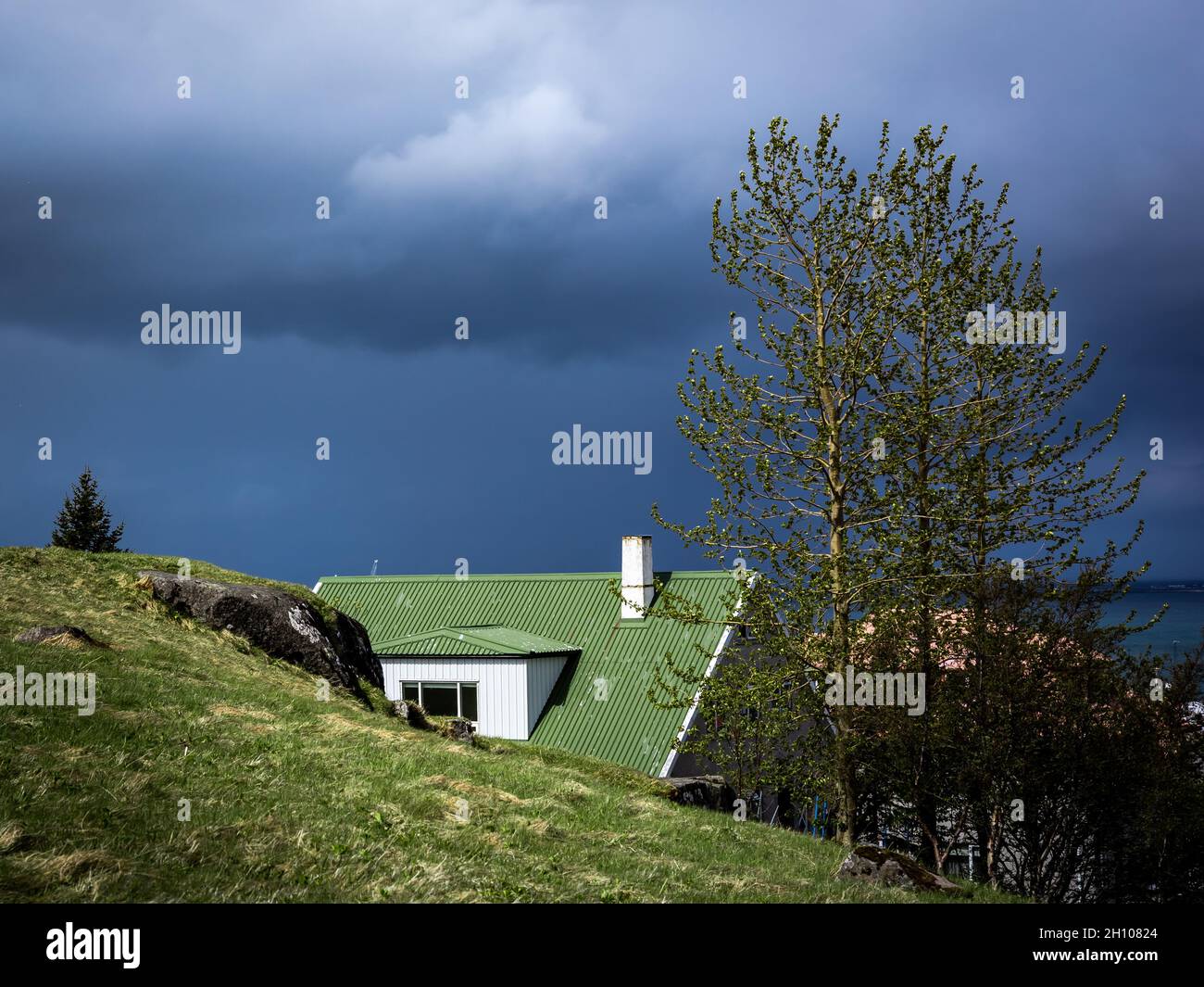 HAFNARFJORDUR, ISLAND - 31. Mai 2021: Ein Haus im skandinavischen Stil mit grünem Dach und Kamin, versteckt hinter grünem Hügel. Dunkler, stürmischer Himmel. Stockfoto