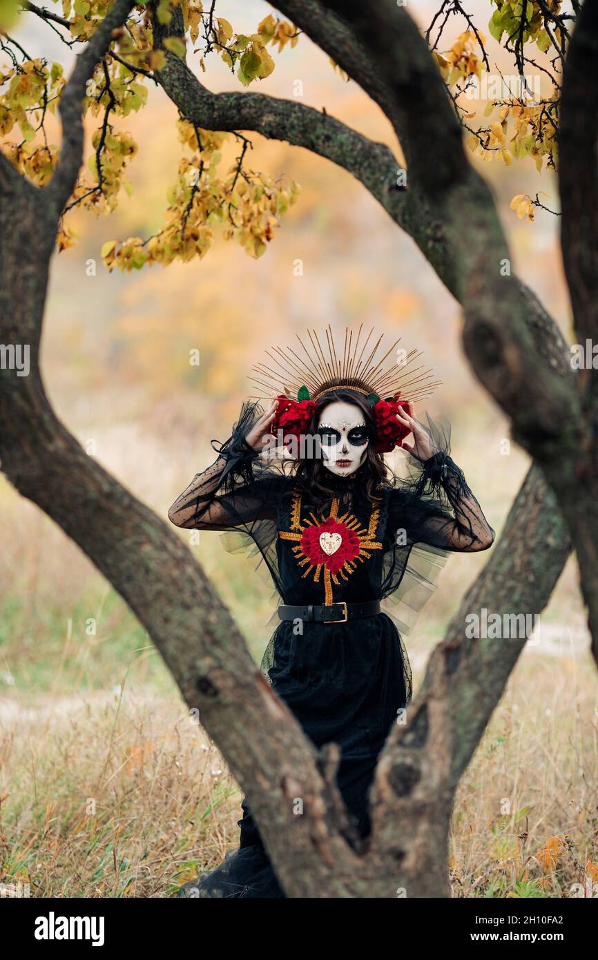 Junge Frau mit Zuckerschädel Make-up und roten Rosen in schwarzem Kostüm des Todes als Santa Muerte ist vor dem Hintergrund des Herbstwaldes gekleidet. Tag des Stockfoto