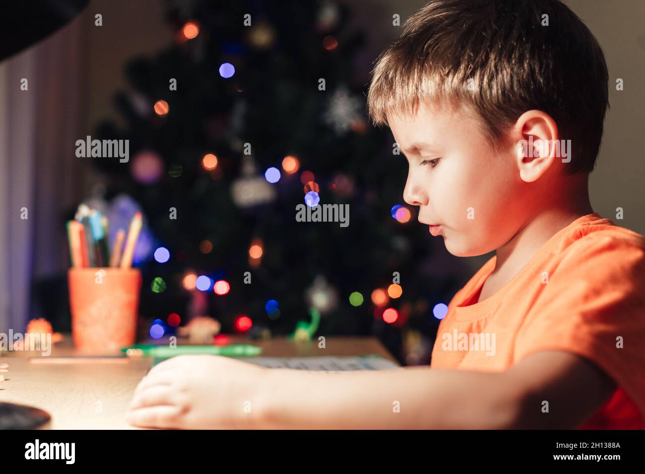 7 Jahre alter Junge, der am Schreibtisch sitzt und Buch liest. Geschmückter Weihnachtsbaum im Hintergrund Stockfoto