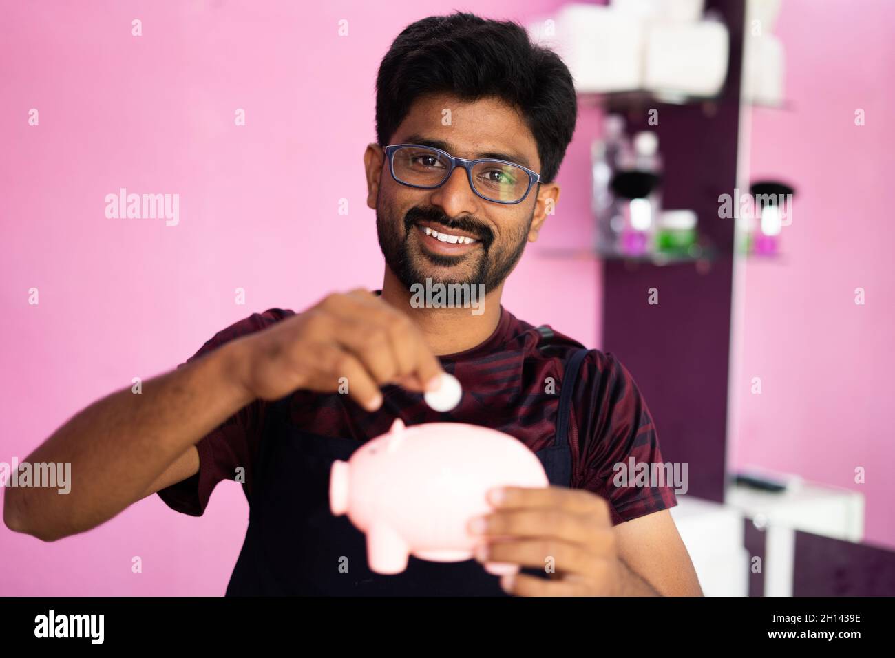 Glücklicher junger professioneller Barbier, der Münzen im Sparschwein platziert, indem er sich die Kamera anschaut - das Konzept der Einsparungen für kleine Unternehmen, Finanzen und Budget. Stockfoto