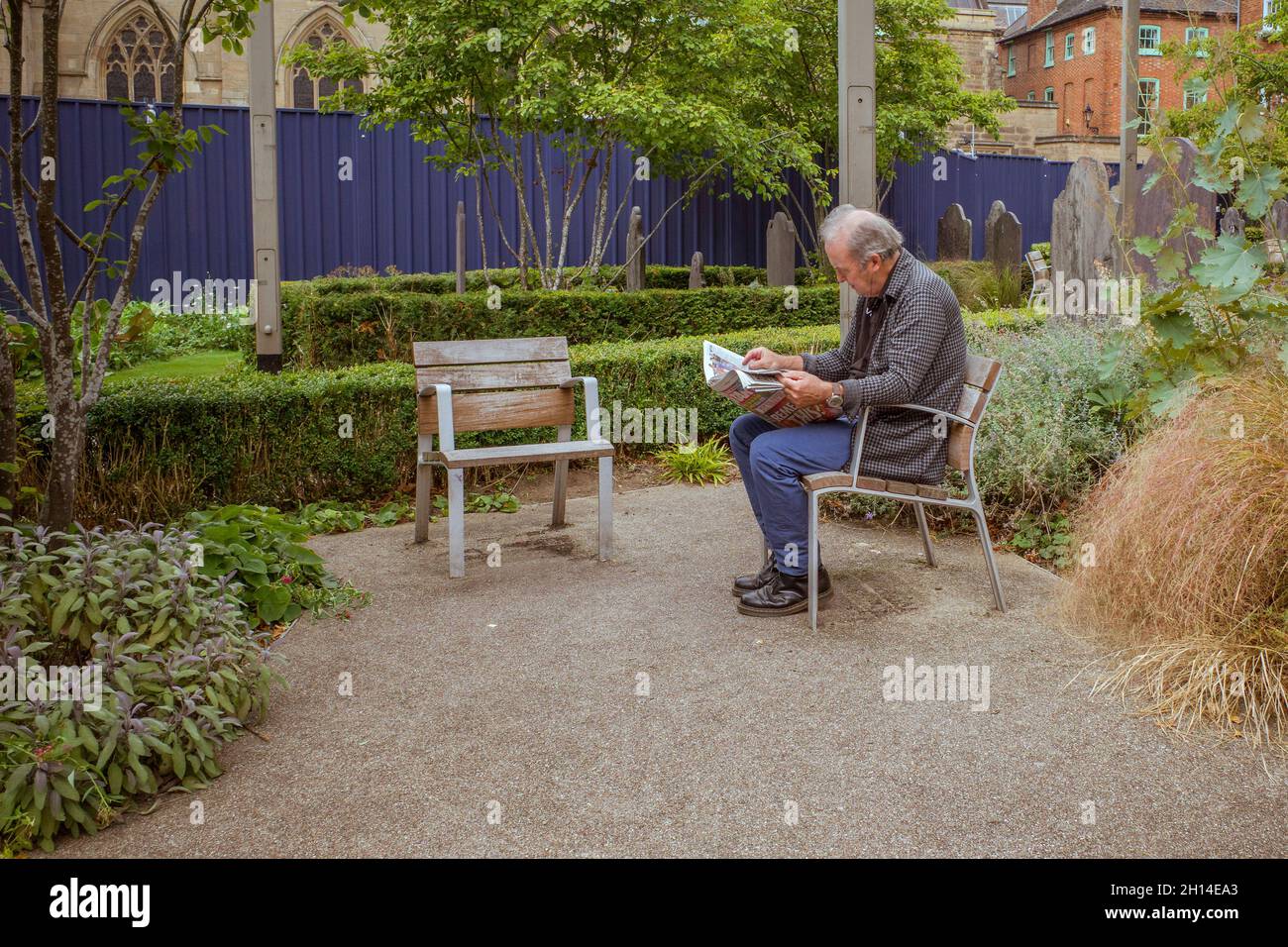 Ein Mann sitzt beim Lesen einer Zeitung in einem öffentlichen Raum neben einem Friedhof. Der Stuhl neben ihm ist konzeptionell ein Raum für abwesende Freunde. Stockfoto