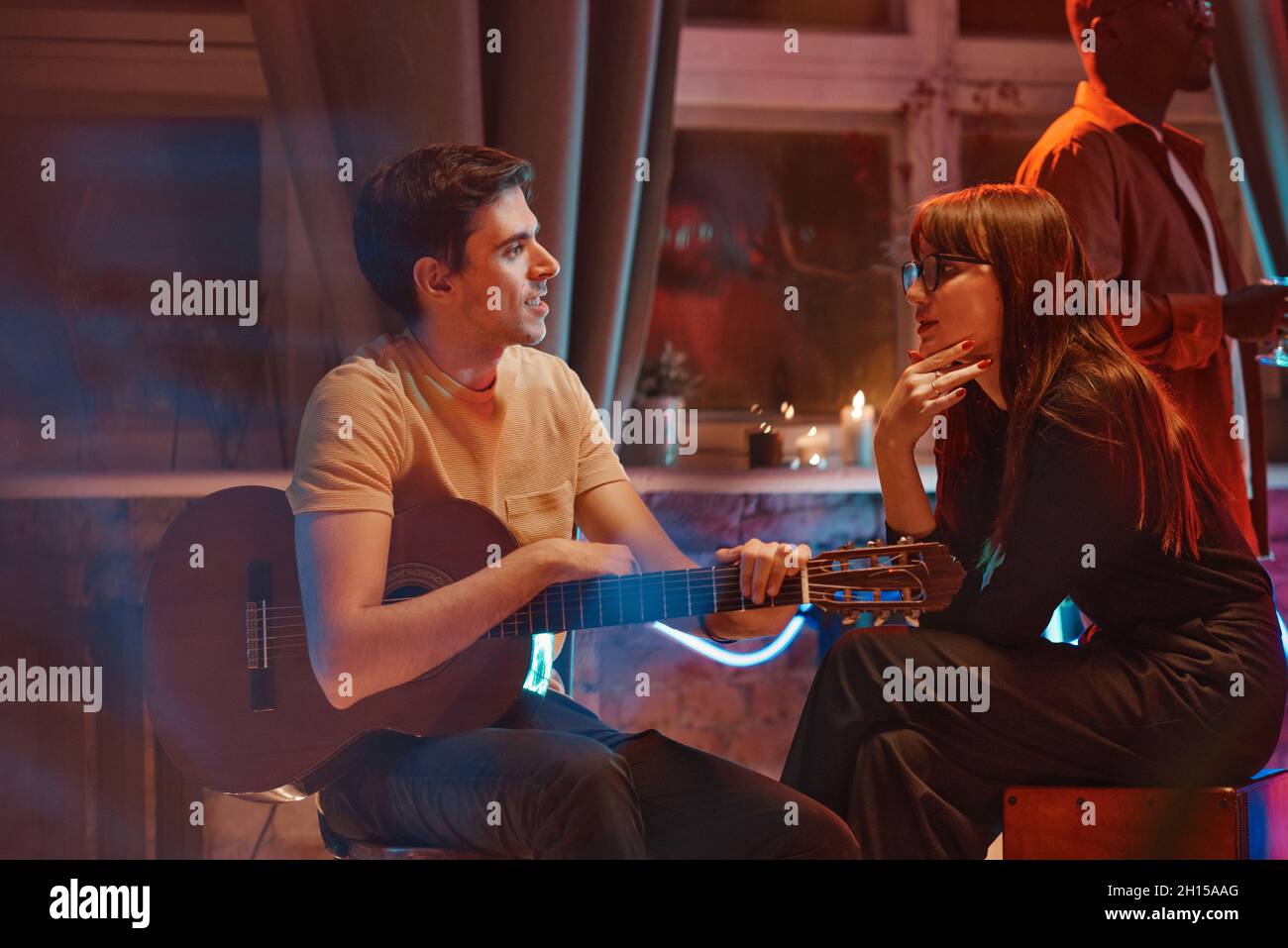 Junge Musikerin mit Gitarre, die mit einer jungen Frau spricht, während sie auf einer Party sitzt Stockfoto