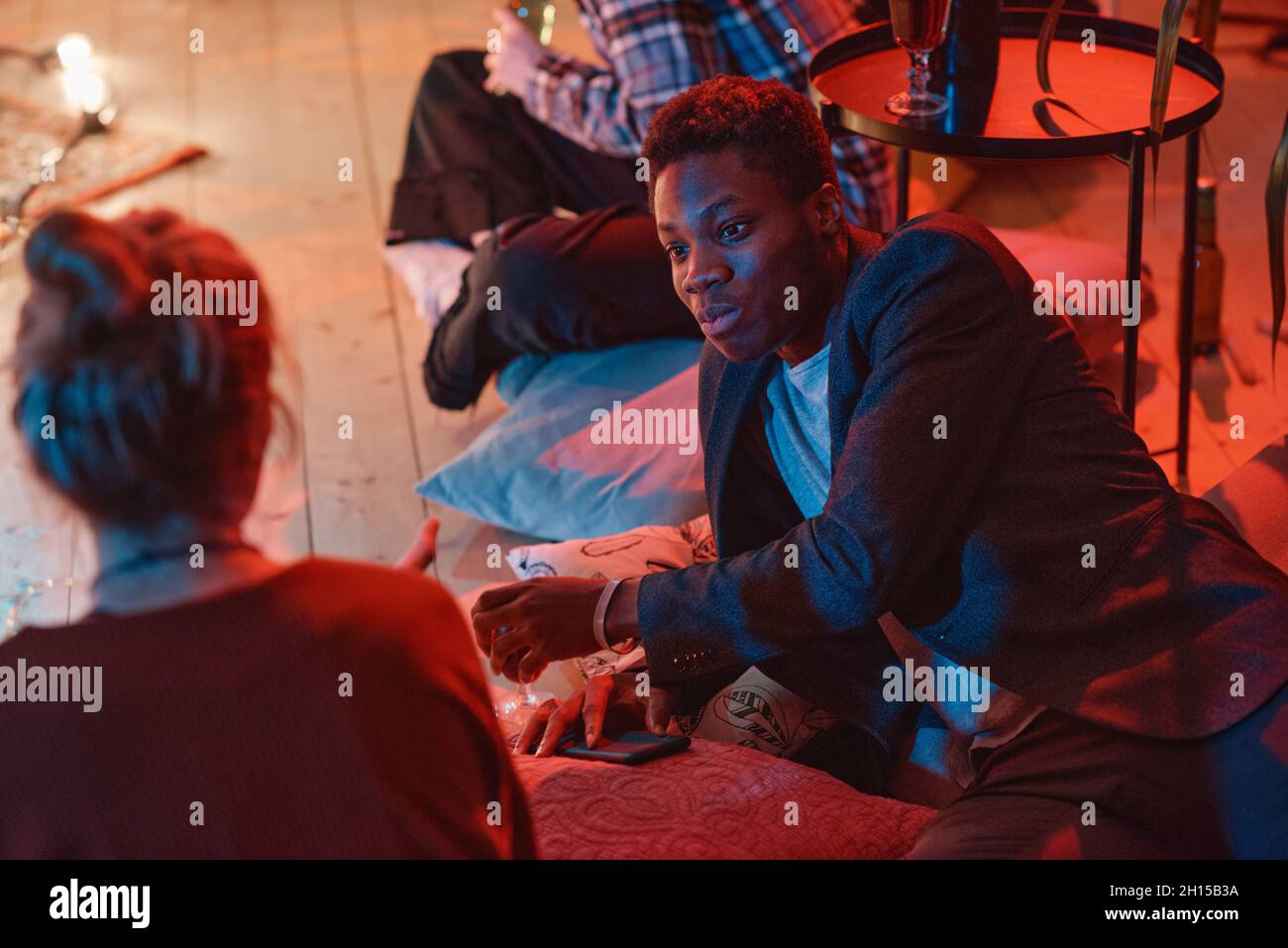 Afrikanischer junger Mann spricht mit einer jungen Frau, während sie auf den Kissen auf dem Boden auf einer Party liegen Stockfoto