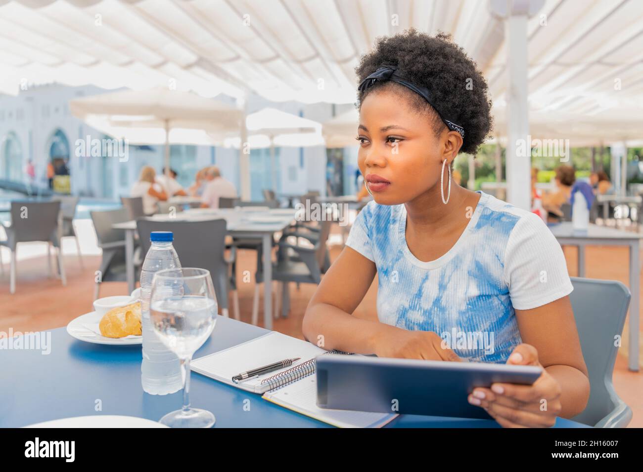 Eine Erwachsene Frau, die mit ihrem digitalen Tablet in einem Freiluftrestaurant mit einem Dach sitzt, während sie mit einem Notizbuch und einem Stift auf dem Tisch wegschaut Stockfoto
