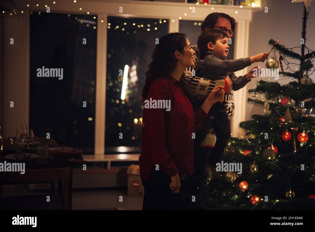 Kleine Familie schmückt Weihnachtsbaum zusammen zu Hause. Eltern mit Kind dekorieren Heim für Weihnachtsfeiern. Stockfoto