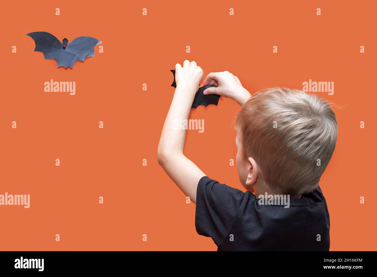 Halloween Kinder. Junge hängende hausgemachte schwarze papiergeschnittene Fledermaus auf orangefarbenem Hintergrund, Kopierbereich, Nahaufnahme. Halloween-Stimmung. Halloween-Heimtextilien Stockfoto