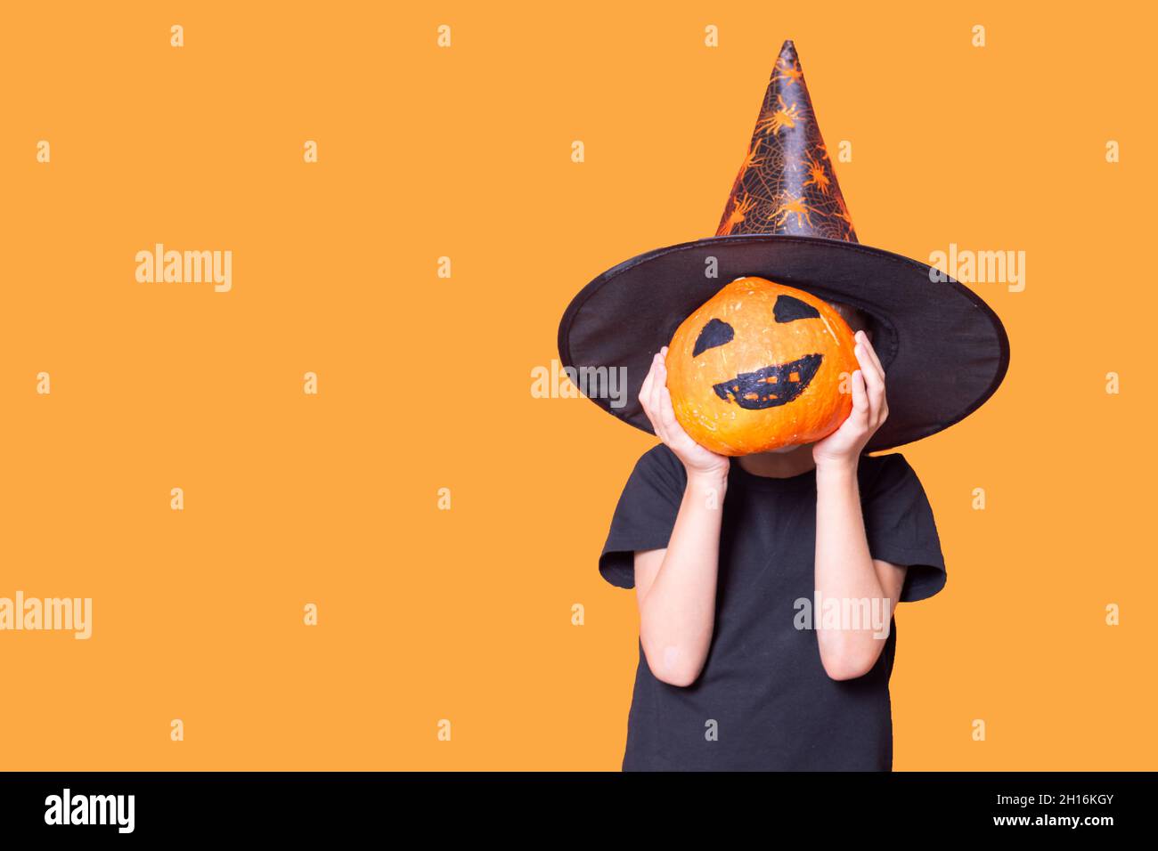 Kinder feiern Halloween. Ein Junge in einem Zauberer Hut hält einen Kürbis vor seinem Gesicht mit einem gemalten gruseligen Gesicht auf einem orangen Hintergrund, c Stockfoto