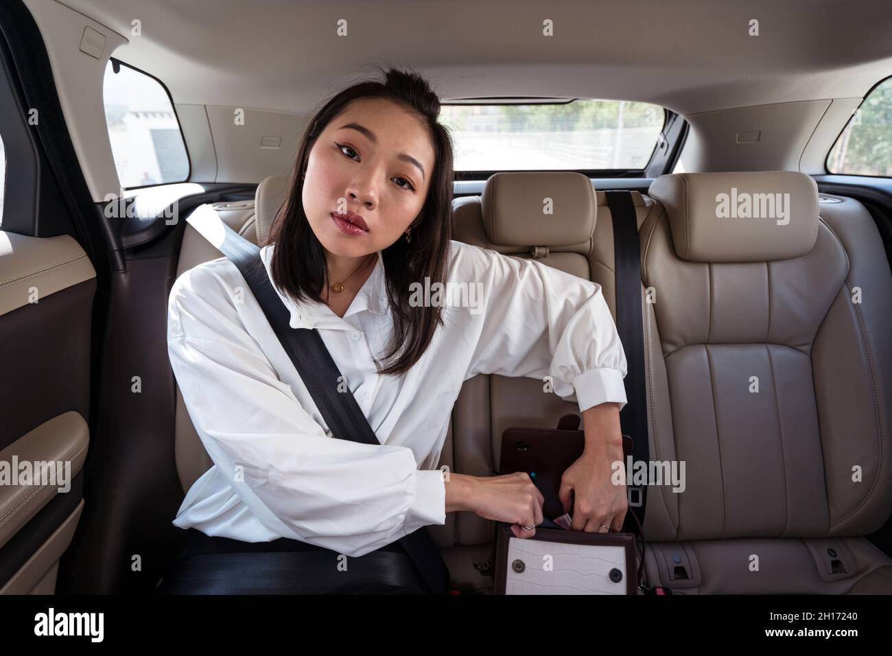 Junge ethnische Frau mit angeschnalltem Sicherheitsgurt, der die Tasche auf dem Rücksitz des Autos öffnet und die Kamera mit Interesse anschaut Stockfoto