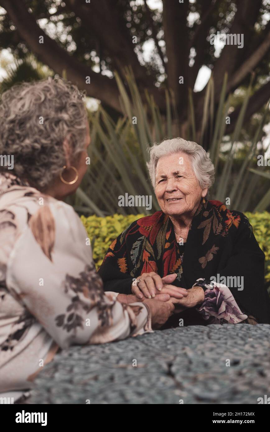 Alte Frau lächelt und hält die Hände, während sie am Tisch im exotischen Hinterhof mit Palmen und Büschen sitzt und sich mit älteren Frauen unterhalten Stockfoto