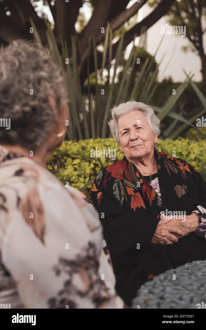 Alte Frau lächelt am Tisch im exotischen Hinterhof mit Palmen und Büschen und genießt das Gespräch mit älteren Freundin Stockfoto