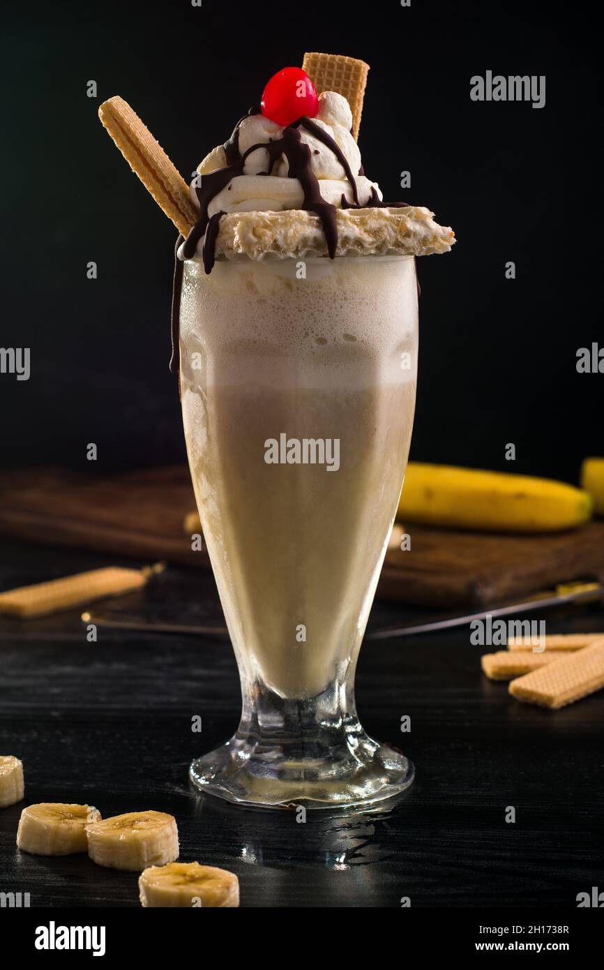 Ein Glas süßer Bananenmilchshake, garniert mit Schlagsahne-Waffeln und Kirsche mit Schokolade darauf Stockfoto
