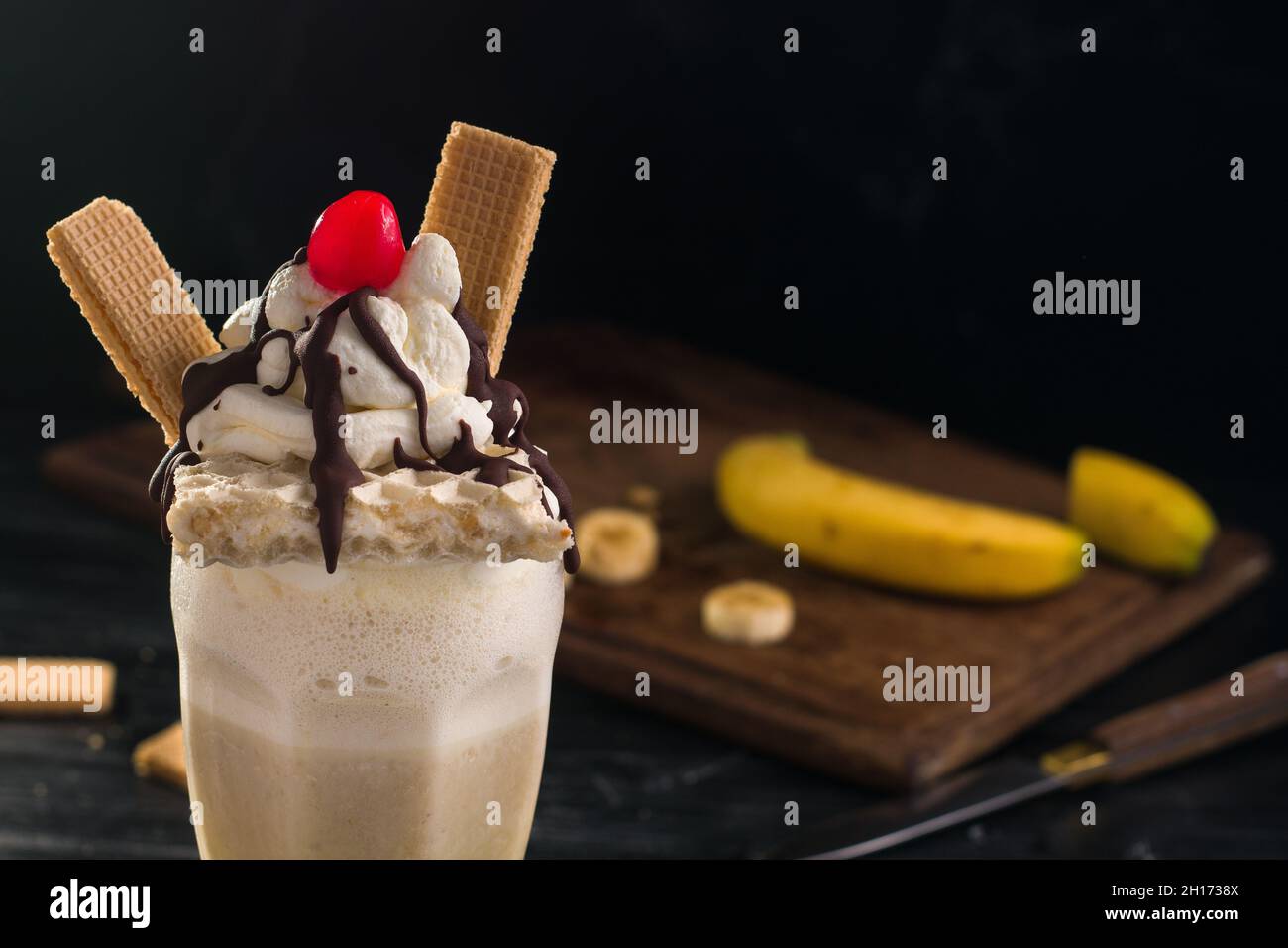 Ein Glas süßer Bananenmilchshake, garniert mit Schlagsahne-Waffeln und Kirsche mit Schokolade darauf Stockfoto