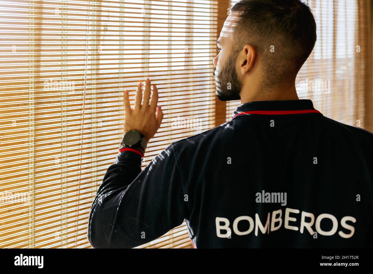 Seitenansicht eines jungen, bärtigen, ethnischen Mannes, der eine Feuerwehrmännerjacke trägt und aus dem Fenster blickt und die Jalousien bei hellem Sonnenlicht mit den Fingern berührt Stockfoto
