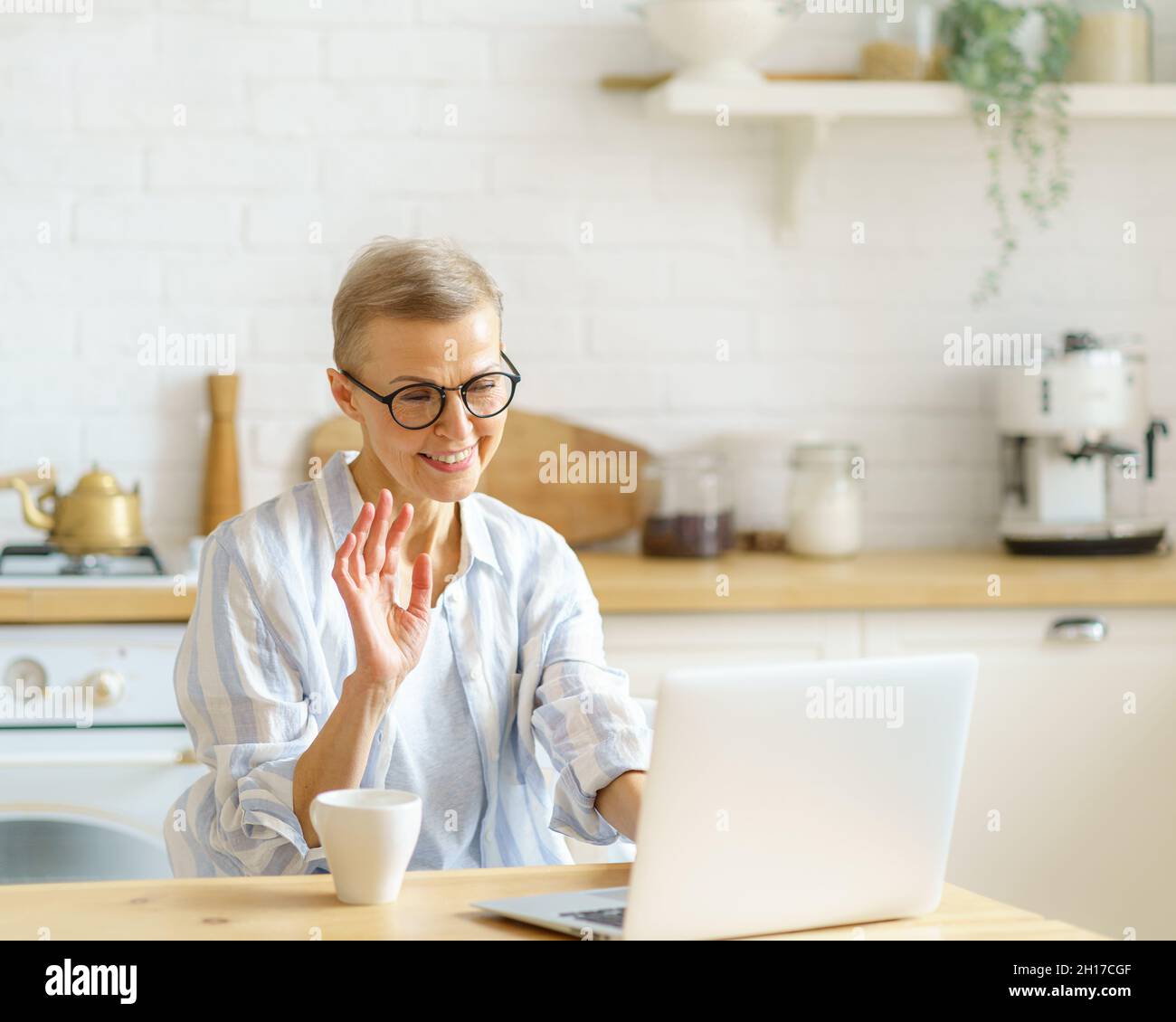 Eine moderne, glückliche ältere Frau mit Brille, die während des Online-Studiums an der Webcam am Laptop winkt Stockfoto
