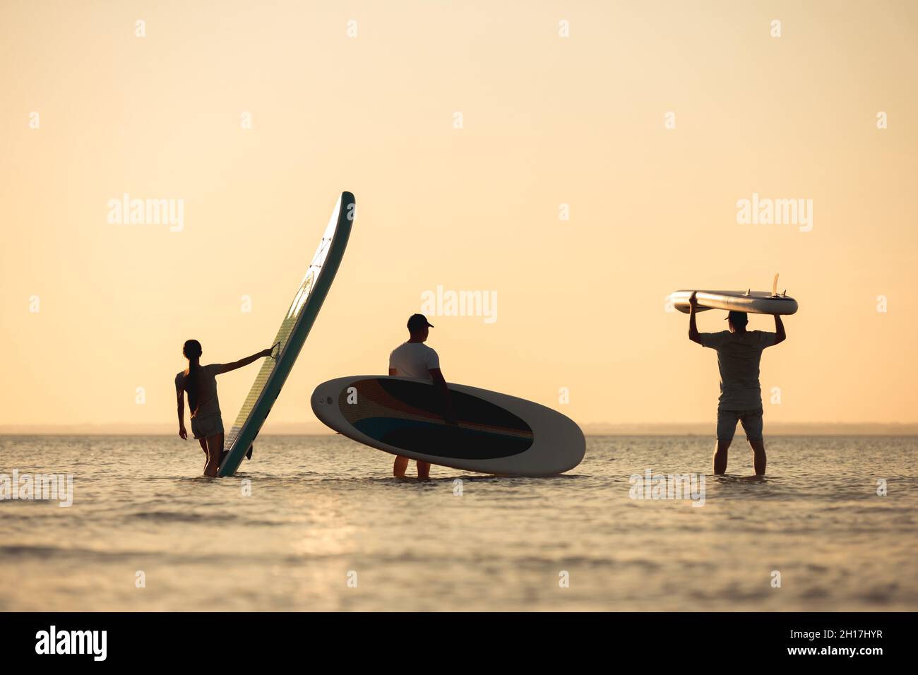 Drei junge Freunde stehen im Wasser mit Brettern in den Händen und schauen auf den Sonnenuntergang Stockfoto