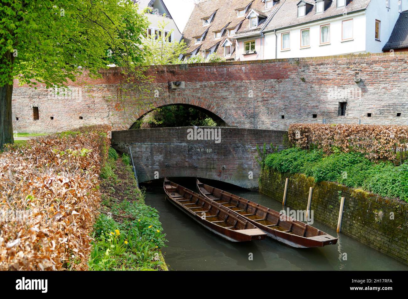 Eine malerische Aussicht auf eine kleine Brücke, darunter Holzboote, grüne Bäume und die alte Stadtmauer in der Stadt Ulm in Deutschland Stockfoto