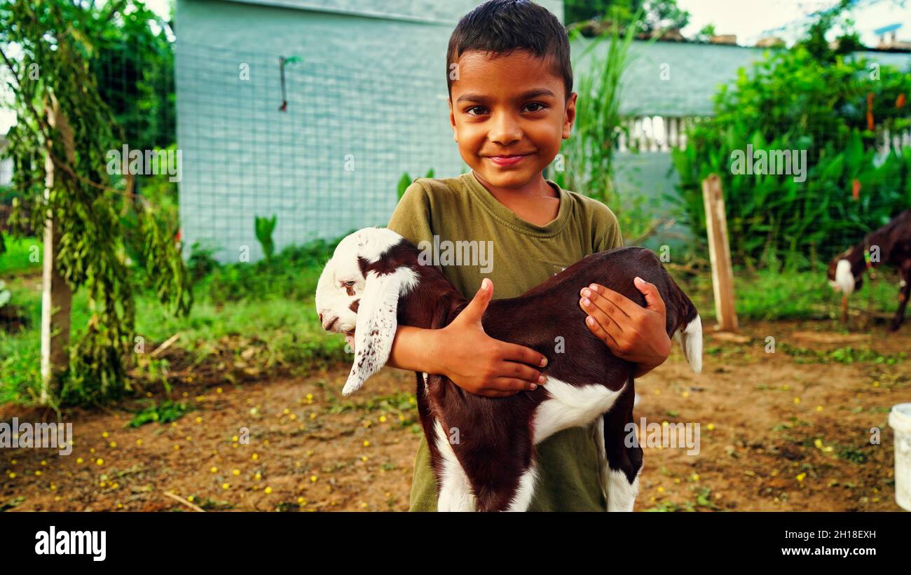 Kleiner indischer Junge, der die kleine Ziege mit Zuneigung und lächelndem Gesicht hält. Stockfoto