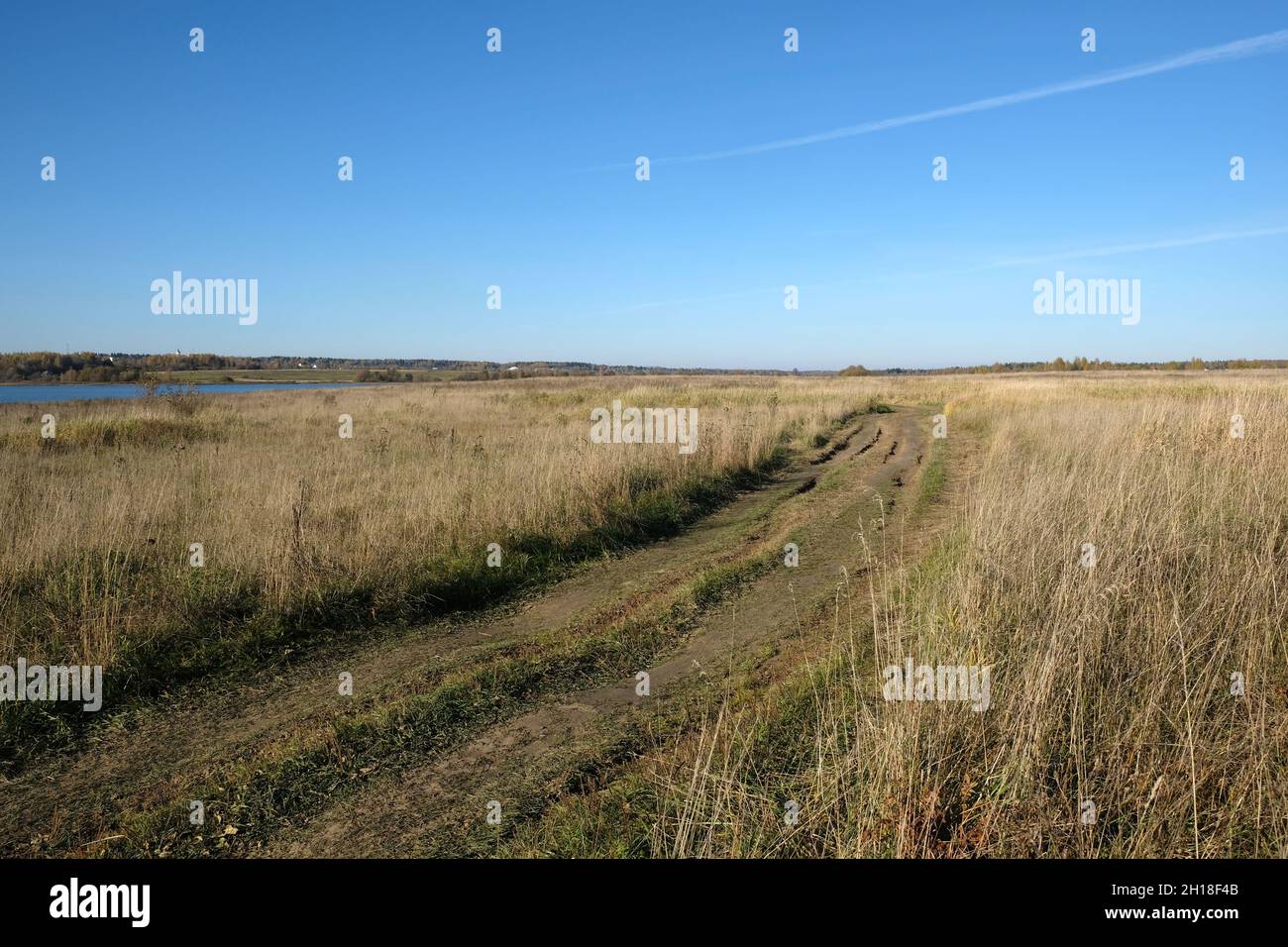Schöne rustikale Landschaft im Frühherbst mit Bodenstraße verläuft durch das Feld in der Nähe von Fluss zu Wald unter klarem blauen Himmel Stockfoto