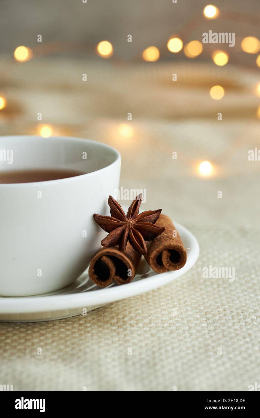 Weiße Tasse Tee mit Zimt auf beigefarbenem Karli in den Weihnachtslichtern. Stillleben zu Weihnachten. Stockfoto