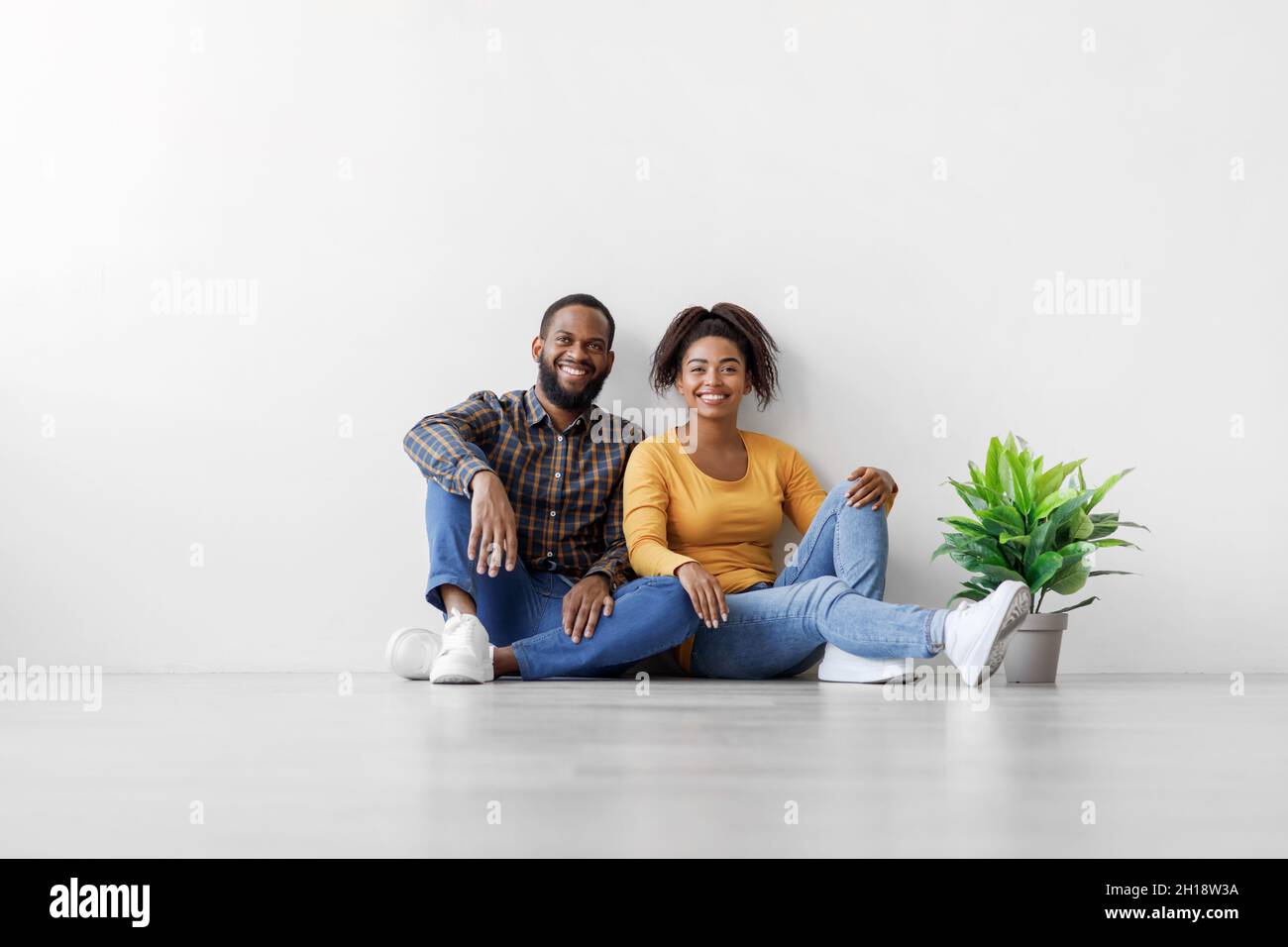 Froher junger afroamerikanischer Mann und Dame sitzen auf dem Boden mit Pflanze im Topf auf weißem Wandhintergrund, ruhen sich nach Umzug aus Stockfoto