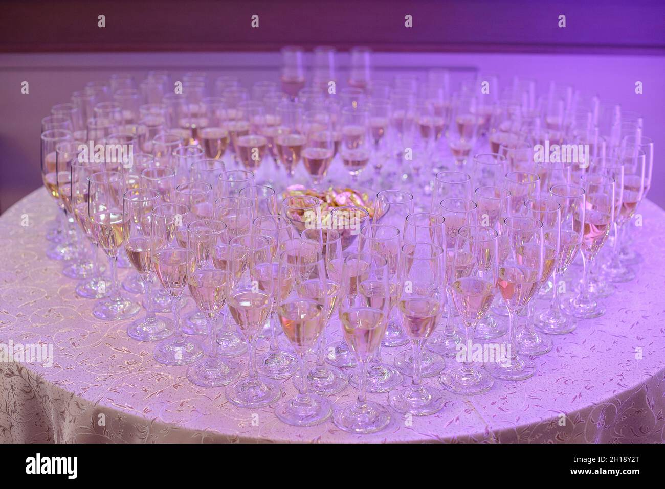 Volle Gläser Sekt oder Champagner bilden eine Herzform, positioniert auf einem runden Tisch unter einem neonvioletten Licht, eine typische Hochzeitsfeier oder Party Stockfoto