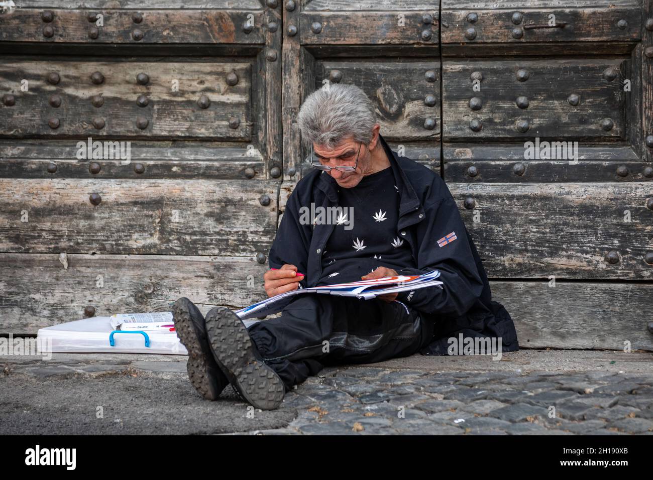 Grauhaariger Mann, der auf dem Boden sitzt, sich an der Tür des Palazzo San Calisto lehnt und von der Piazza di Santa Maria in Trastevere, Rom, Italien, skizziert Stockfoto