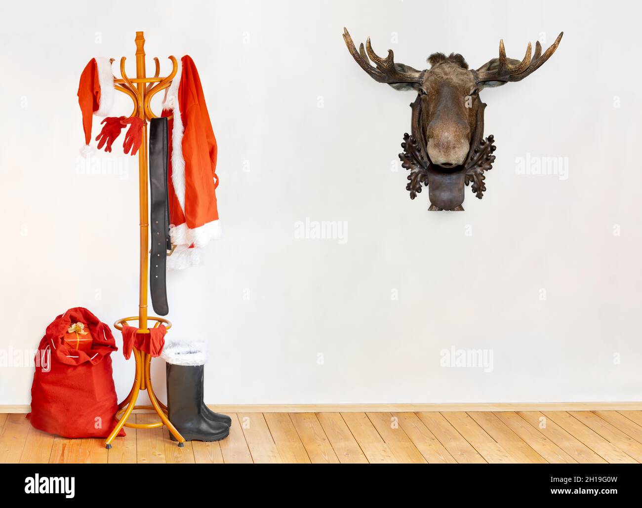 Die Ausrüstung des Weihnachtsmannes hängt am Kleiderhaken in einem Raum, wobei der Kopf des Rentieres als Trophäe an einer weißen Wand hängt. Stockfoto