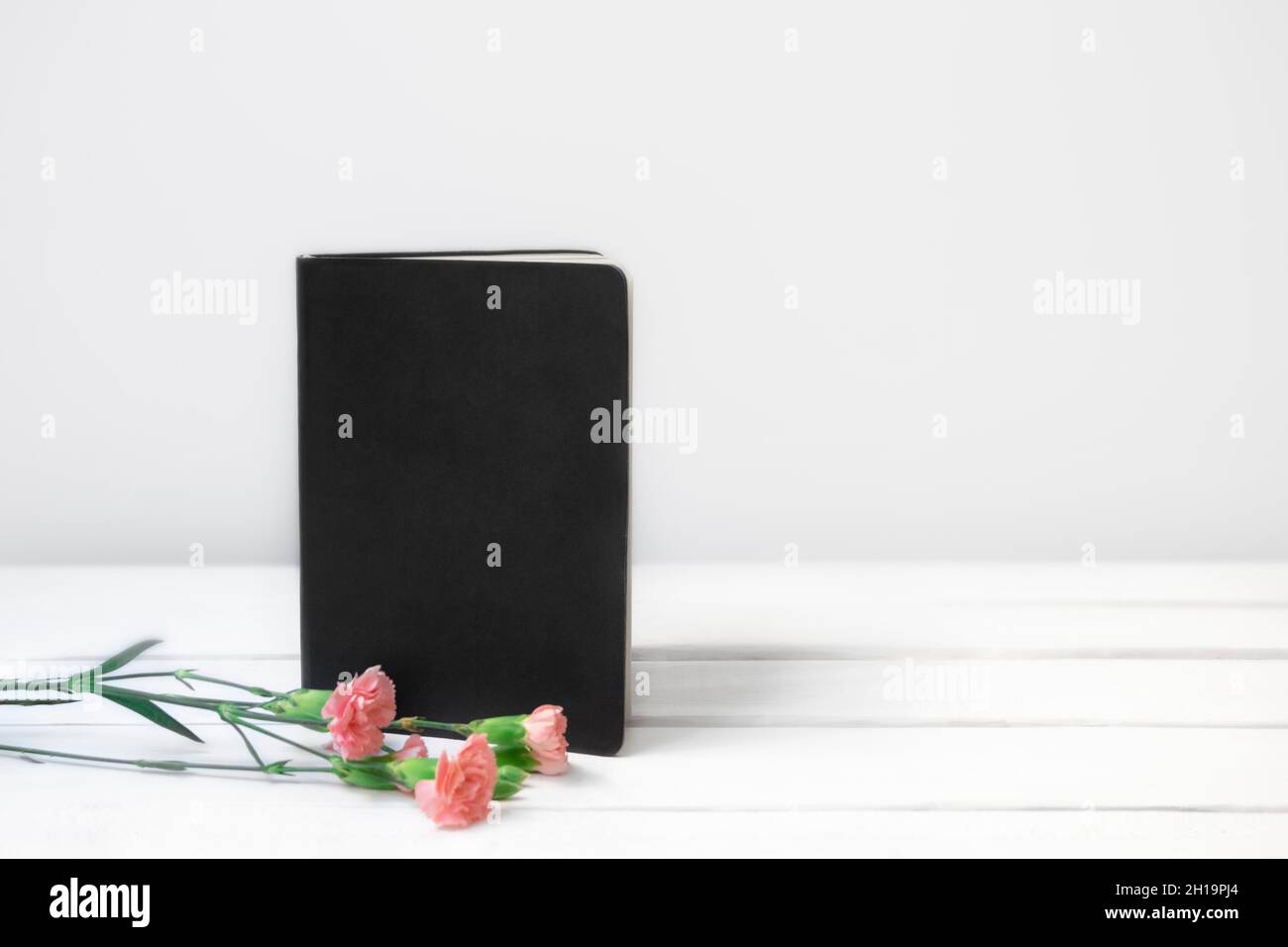 Weißer Holzschreibtisch mit schwarzem leerem Buch oder Notizbuch und rosa Blumen. Vorderansicht. Platz für Text, Platz zum Kopieren Stockfoto
