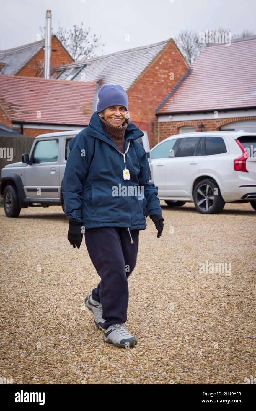Ältere asiatische Inderin, die lächelt und mit einem persönlichen Sicherheitsalarm nach draußen geht. Winter in Großbritannien Stockfoto