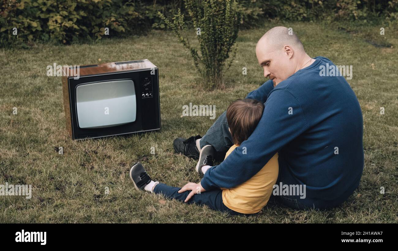 Der Mann, Vater mit einem kleinen Kind, sitzt auf dem Gras und schaut sich einen alten Retro-Fernseher an Stockfoto