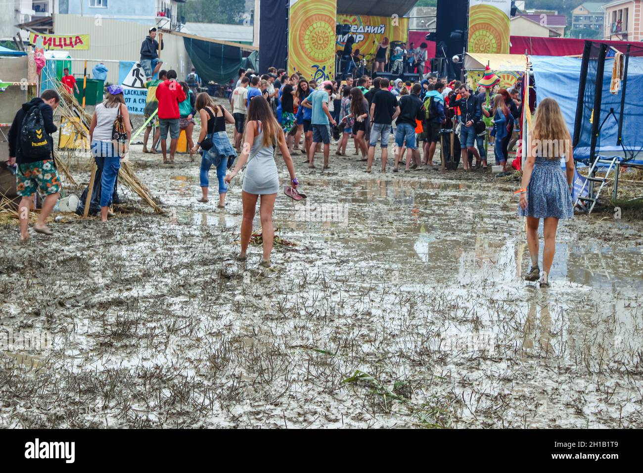 Mädchen, die auf Schlamm laufen. Im Hintergrund ist eine Musikszene und Menschen. Schlechtes Wetter während des Musikfestivals. Republik Krim, Sudak, 27. Juli 2013 Stockfoto