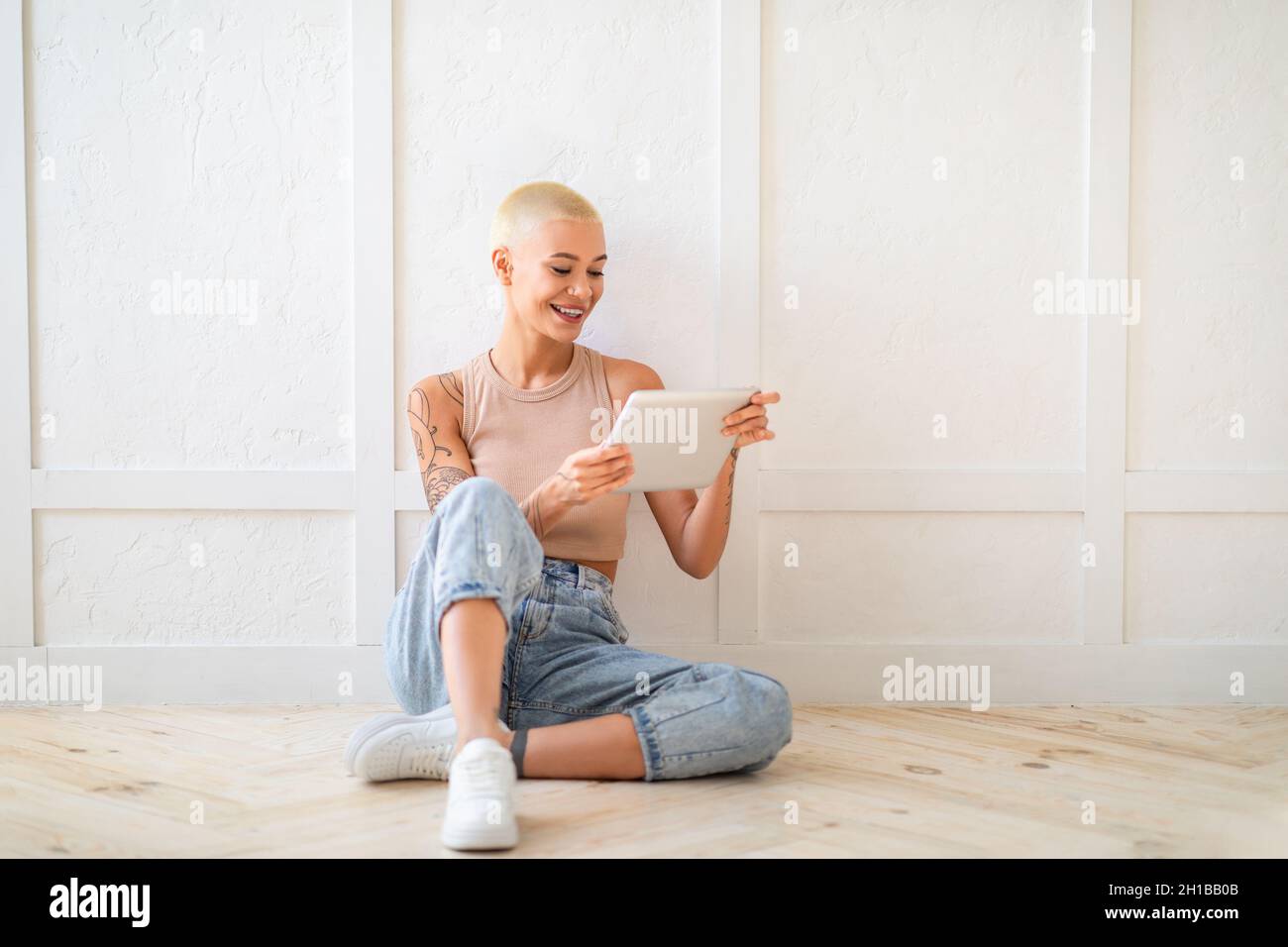 Glückliche junge Frau, die ein digitales Tablet nutzt, im Internet surft oder mit Freunden chattet, auf dem Boden über einer weißen Wand sitzt Stockfoto