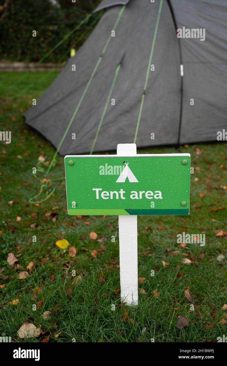 Grün-weißes Zeltareal mit Text und Symbol. Verschwommener Hintergrund aus Zelt und Gras mit Herbstblättern. Regentropfen auf Schild. Keine Personen. Stockfoto