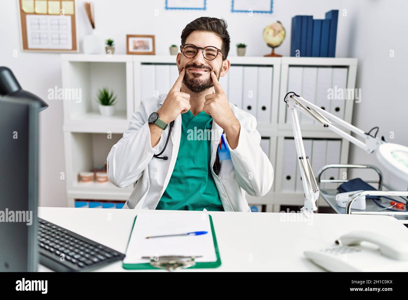 Junger Mann mit Bart, der eine Arztuniform und ein Stethoskop in der Klinik trug, lächelte mit offenem Mund, die Finger zeigten und ein fröhliches Lächeln erzwangen Stockfoto