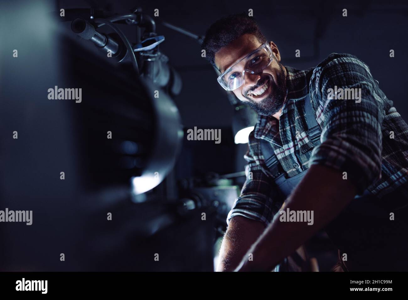 Junger afroamerikanischer Industriemann während der Nachtschicht in Innenräumen in einer Metallwerkstatt, der die Kamera ansah Stockfoto