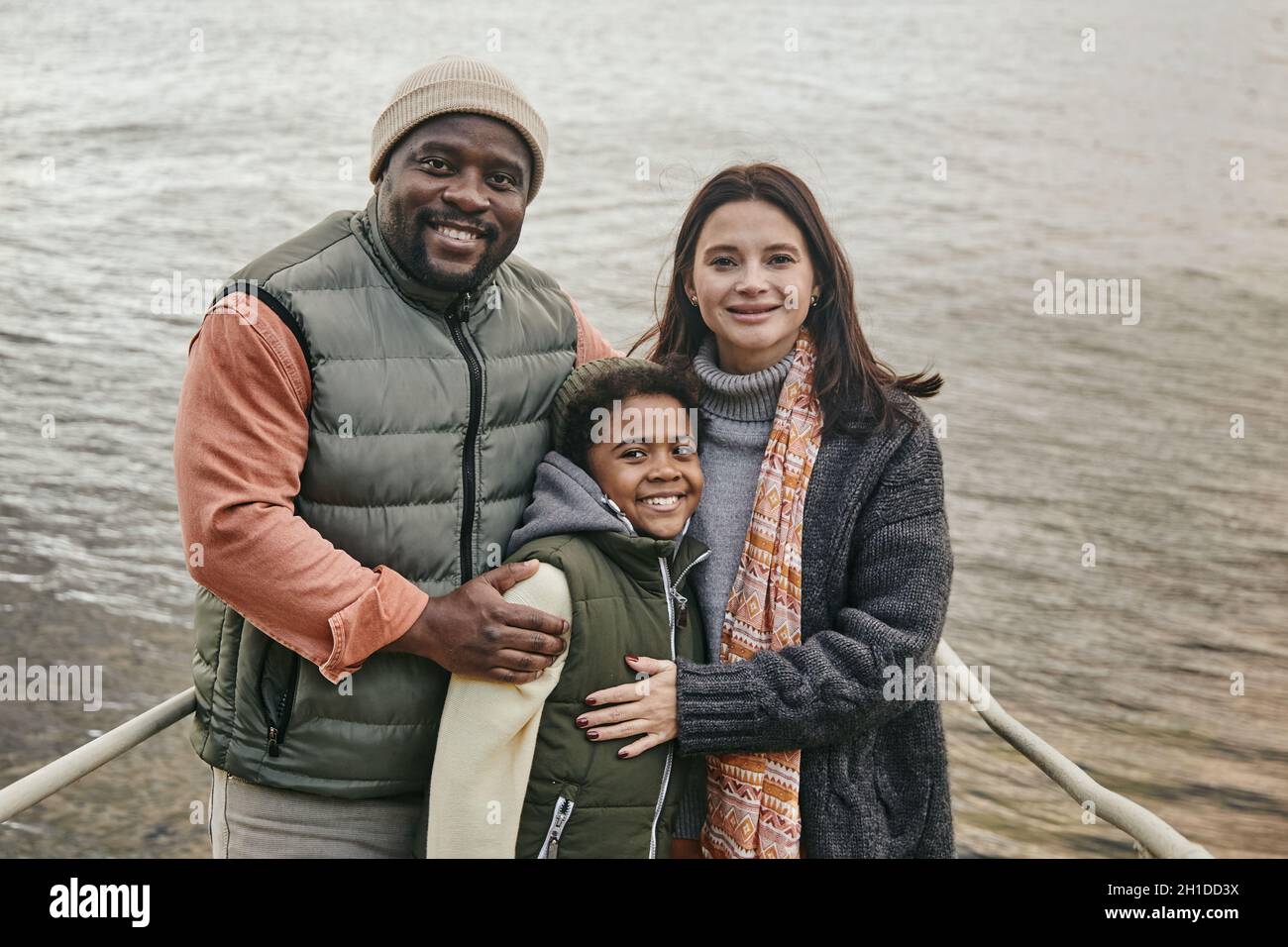 Porträt einer multiethnischen Familie, die ihren Sohn umarmt und die Kamera anlächelt, während sie im Freien gegen den See steht Stockfoto
