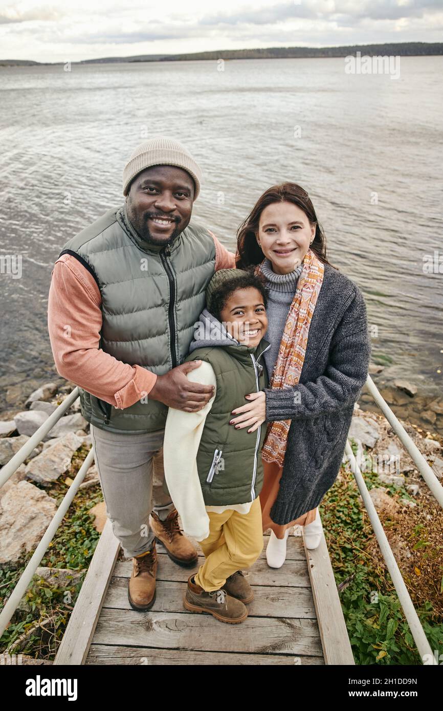 Porträt einer glücklichen, multiethnischen Familie von drei lächelnden Kindern, die auf der Brücke gegen den See stehen Stockfoto