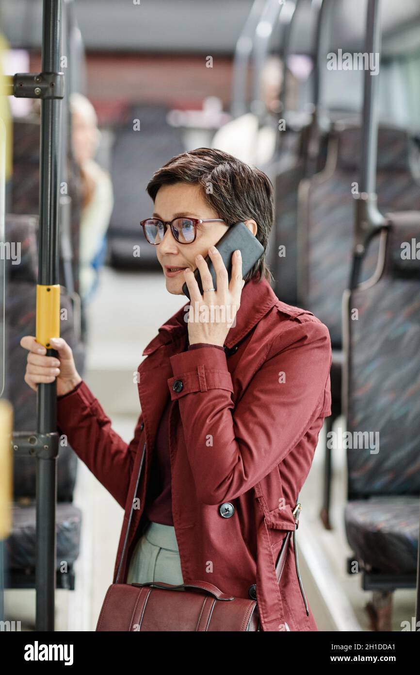 Vertikales Porträt einer eleganten erwachsenen Frau, die mit dem Smartphone im Bus spricht, während sie mit öffentlichen Verkehrsmitteln in der Stadt unterwegs ist Stockfoto