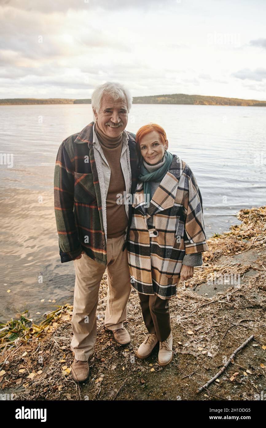 Porträt eines glücklichen älteren Ehepaares, das die Kamera umarmt und lächelt, während es im Freien in der Nähe des Sees steht Stockfoto