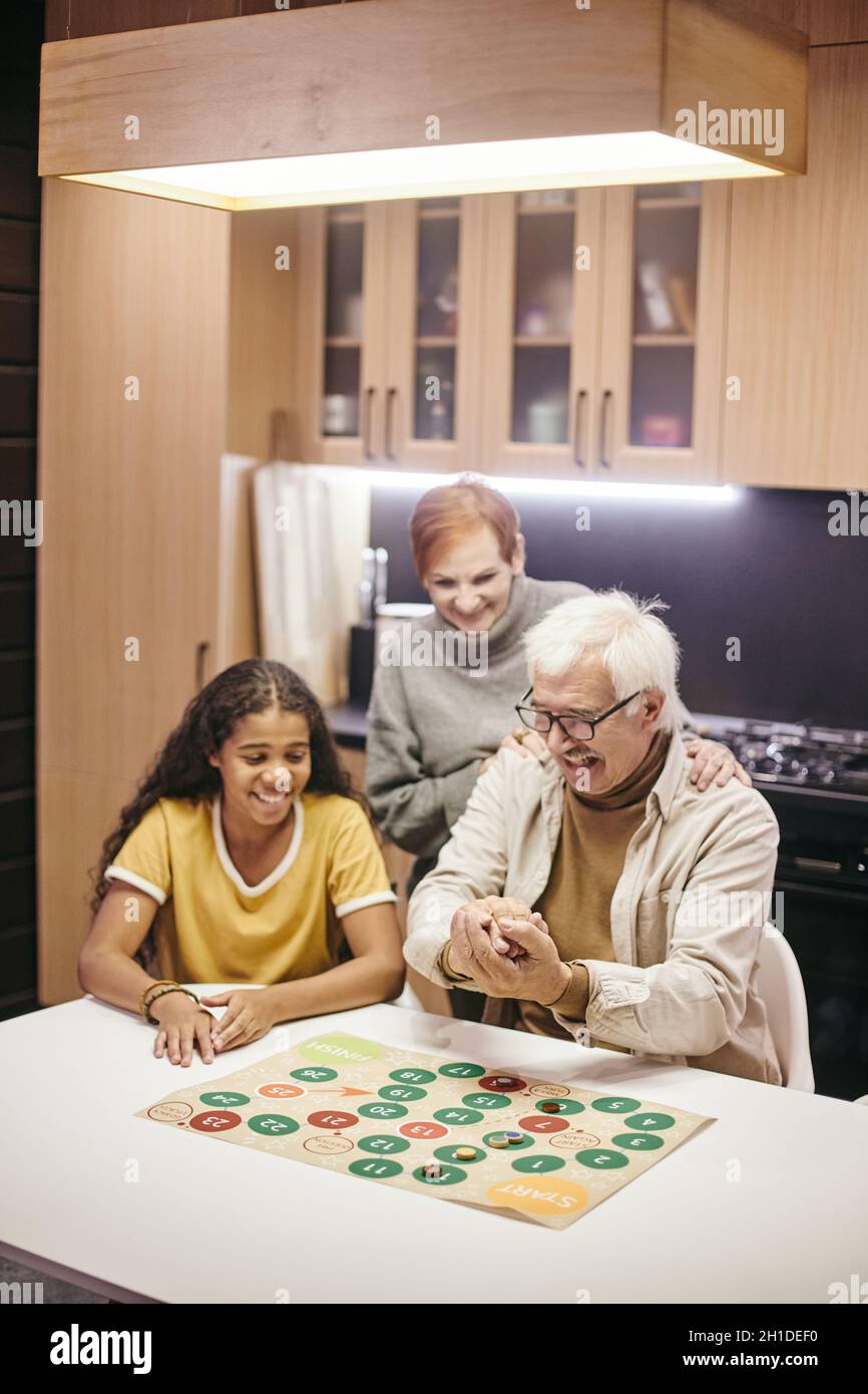 Glückliche Großeltern sitzen am Tisch in der Küche und spielen Brettspiel mit ihrer Enkelin Stockfoto