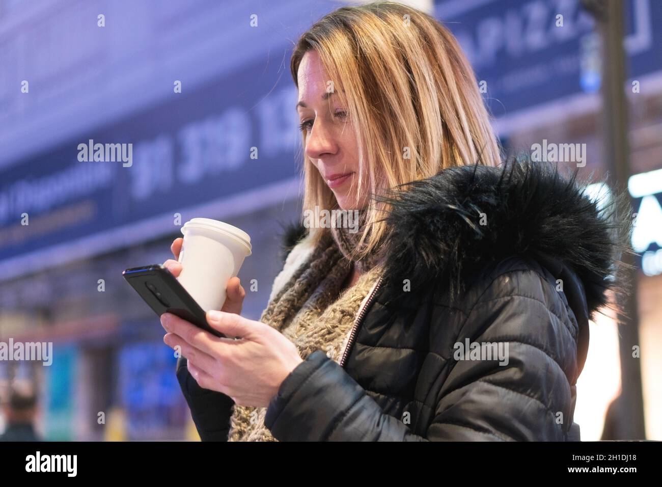 Hübsche, junge Frau mit Smartphone und Kaffee Lächeln glücklich in der Stadt bei Nacht. Stockfoto